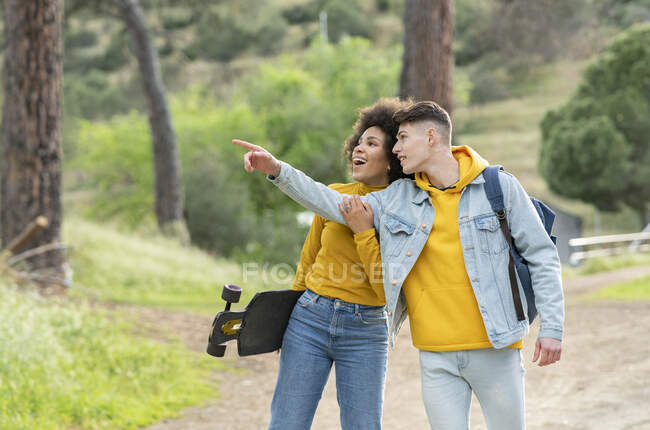 Diverso joven hombre y mujer con longboard tomados de la mano y caminando por el camino del campo en verano día de fin de semana mirando hacia otro lado - foto de stock