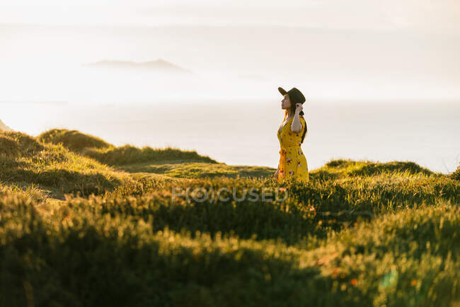 Jolie jeune femelle en robe jaune et chapeau debout sur une prairie herbeuse verdoyante dans une campagne ensoleillée — Photo de stock