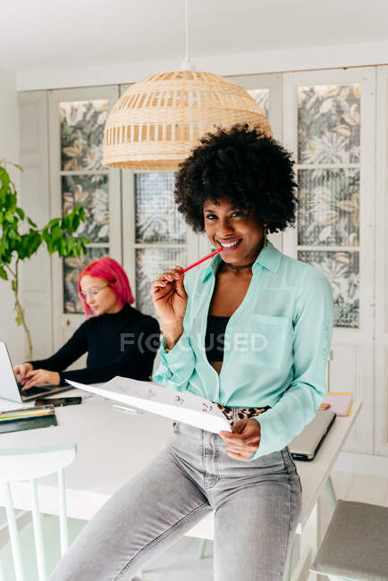 Moderna donna afro-americana freelance di successo in abito elegante con capelli afro sorridente guardando la fotocamera mentre si siede a tavola e leggere il documento a casa — Foto stock