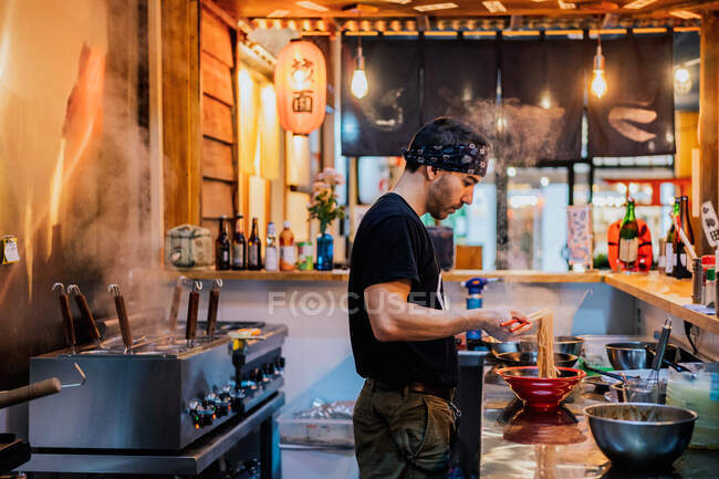 Vista lateral del hombre en bandana de pie en el mostrador y ramen de cocina en la cafetería asiática moderna - foto de stock
