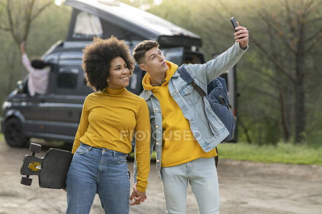 Diverso joven hombre y mujer con monopatín tomando selfie cerca de furgoneta durante el viaje por carretera en el día de verano en la naturaleza - foto de stock