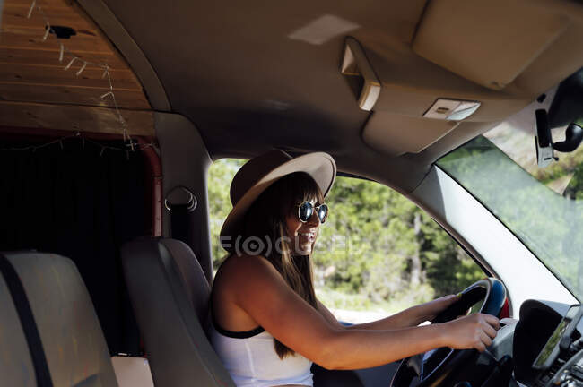 Vista lateral de la viajera sentada en el asiento del conductor en furgoneta y disfrutando del viaje por carretera en verano - foto de stock