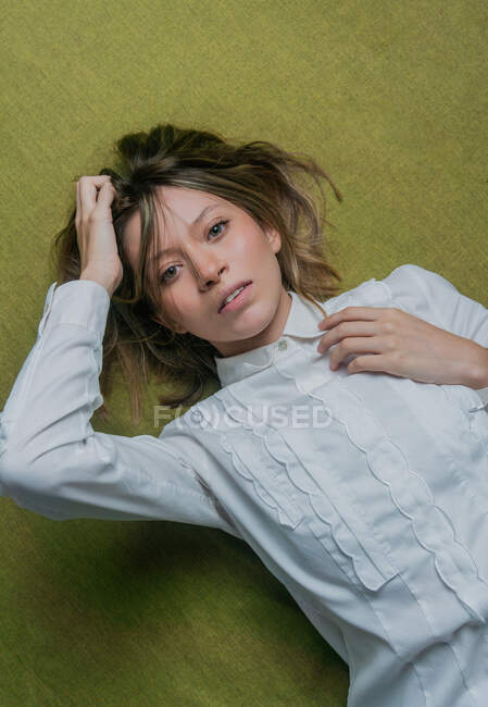 From above of young green eyed female with long fair hair wearing white blouse touching head and looking at camera while lying on green fabric — Stock Photo