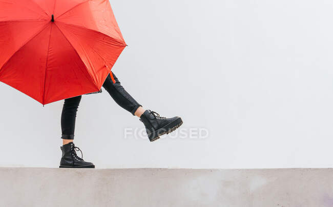 Anonimo giovane femmina con ombrello rosso che cammina e si equilibra sul confine contro il muro grigio nella giornata piovosa sulla strada — Foto stock