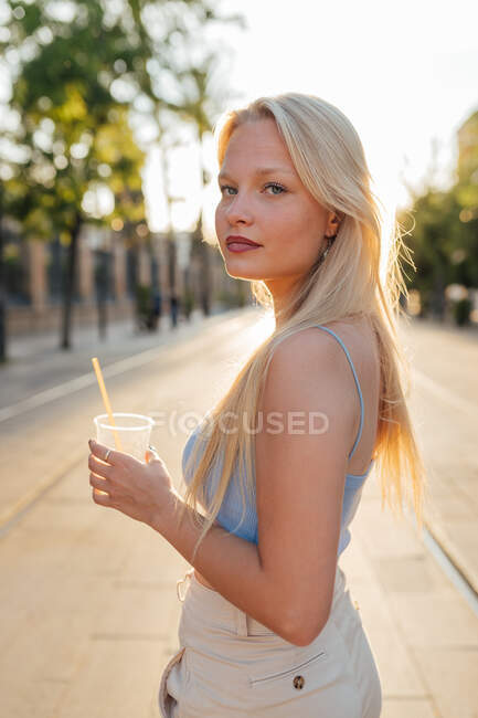 Vista lateral da fêmea de pé com limonada fria em copo de plástico na rua no verão olhando para a câmera — Fotografia de Stock