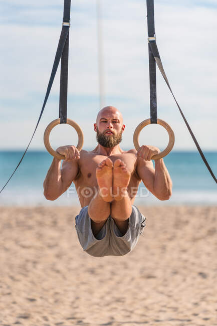 Мужчина без рубашки с бородой висит на гимнастических кольцах с поднятыми ногами, упорно тренируясь на песчаном пляже, отворачиваясь — стоковое фото