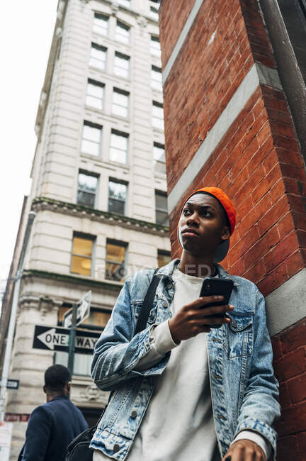 From below thoughtful African American guy in denim trendy jacket and cap surfing modern mobile phone during city stroll — Stock Photo
