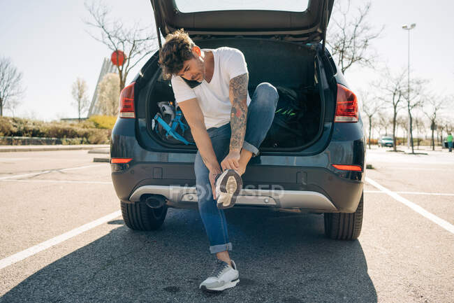 Athlète masculin tatoué portant des chaussures tout en parlant sur un téléphone portable contre une voiture avec coffre ouvert en ville — Photo de stock
