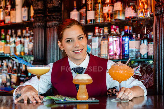 Smiling female barkeeper standing at bar counter with different types of alcohol drinks served in creative cocktail glasses in shape of mushroom and fish — Stock Photo