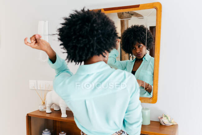 Vue arrière d'une belle jeune femme afro-américaine en tenue décontractée touchant les cheveux bouclés tout en se tenant dans la chambre et en regardant le miroir — Photo de stock