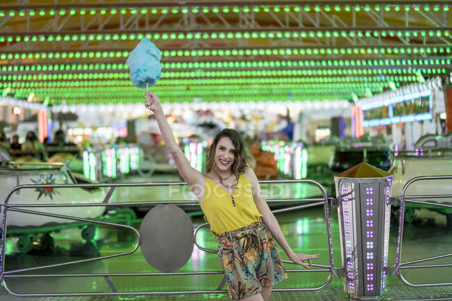 Оптимістична жінка стоїть з блакитною бавовняною цукеркою в піднятій руці, насолоджуючись вихідними в парку розваг і дивлячись на камеру — стокове фото