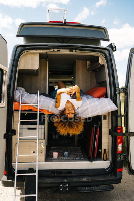 Positive schwarze Frau mit Afro-Frisur liegt kopfüber auf dem Bett im Wohnwagen und surft an sonnigen Sommertagen am Handy — Stockfoto