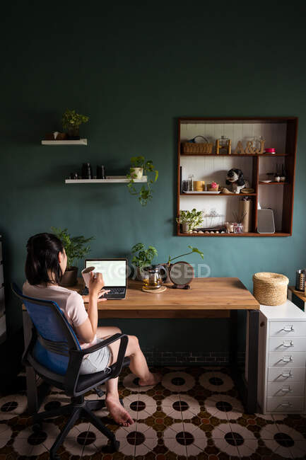 Vista lateral de la mujer asiática freelancer leer documentos en el ordenador portátil mientras está sentado y beber té en la mesa durante el trabajo remoto - foto de stock