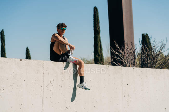 Спортсмен в активному одязі з пляшкою води після тренування проти дерев на сонячному світлі — стокове фото