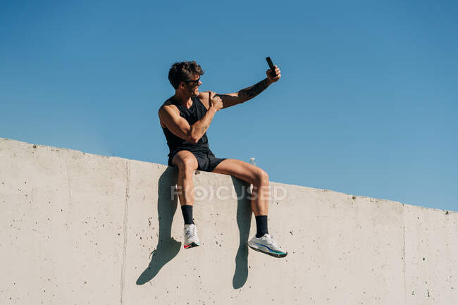 Fit male athlete in sports clothes taking selfie with cellphone while resting on fence after workout — Stock Photo