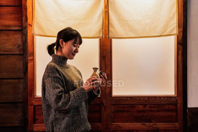 Side view of overjoyed Asian woman in sweater standing near door with traditional drink in cafe — Stock Photo