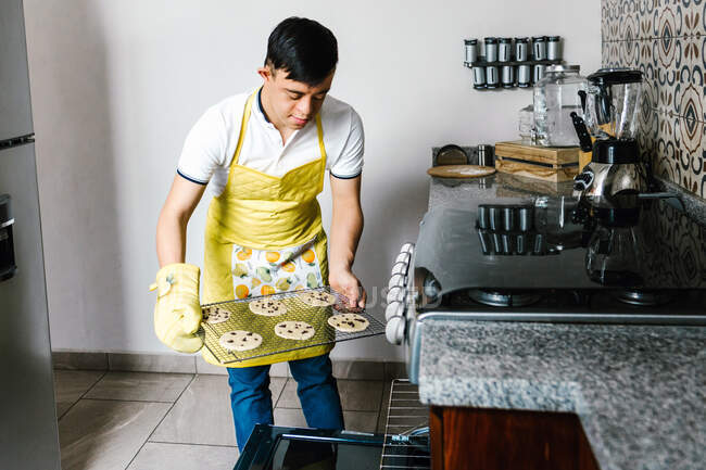 Adolescente étnico con síndrome de Down poniendo galletas de chocolate crudo en el horno mientras hornea pastelería en la cocina - foto de stock