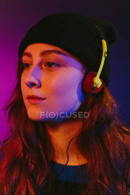 Young stylish female millennial in retro inspired outfit and hat listening to music in headphones using vintage tape cassette player against purple background looking away — Stock Photo