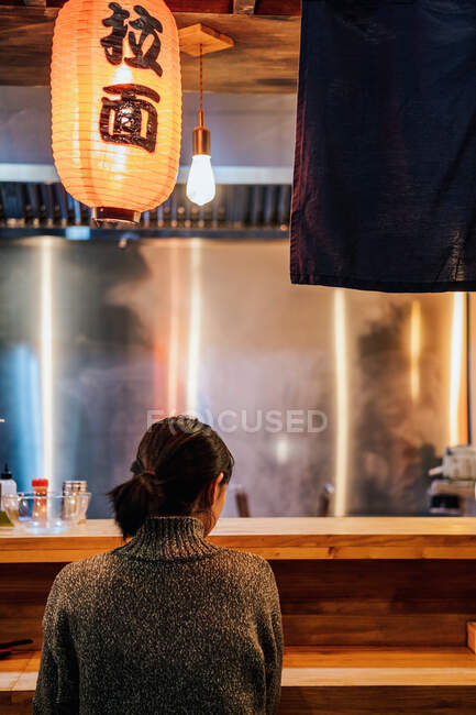 Vista posterior de la mujer de pelo negro sin rostro en suéter sentado en el mostrador en el acogedor bar ramen - foto de stock