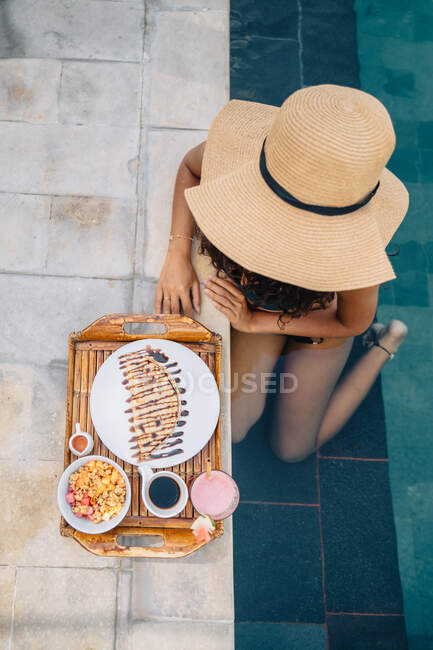 Desde arriba turista femenina anónima apoyada en la piscina contra bandeja con delicioso desayuno a la luz del sol - foto de stock