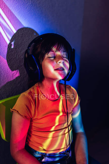 Chica feliz en ropa casual tableta de navegación y escuchar canciones favoritas con los ojos cerrados a través de auriculares mientras está sentado en luces de neón - foto de stock