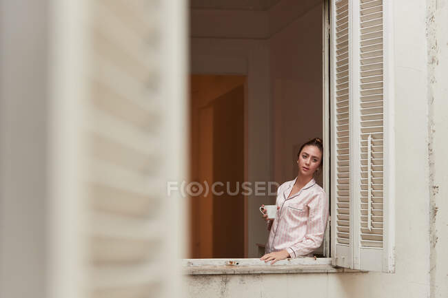 Tranquilo fêmea no pijama de pé perto da janela com xícara de café da manhã e olhando para longe — Fotografia de Stock