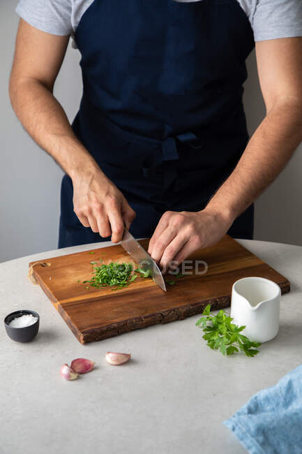 Неузнаваемый мужчина в фартуке режет свежую петрушку на деревянной доске возле соли и чеснока во время обеда дома — стоковое фото