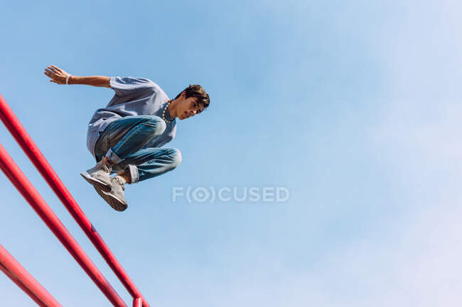 Снизу храбрый мужчина перепрыгивает через металлический забор на улице и показывает паркур трюк против голубого неба — стоковое фото