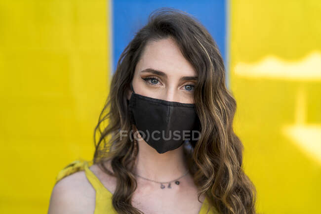 Conteúdo fêmea com cabelo ondulado usando máscara protetora durante coronavírus na cidade olhando para a câmera em dois fundos coloridos — Fotografia de Stock