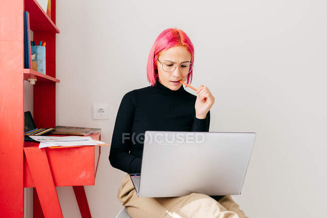 Joven freelancer enfocada en ropa casual sentada en silla y usando laptop mientras trabaja en proyecto en apartamento moderno y ligero - foto de stock
