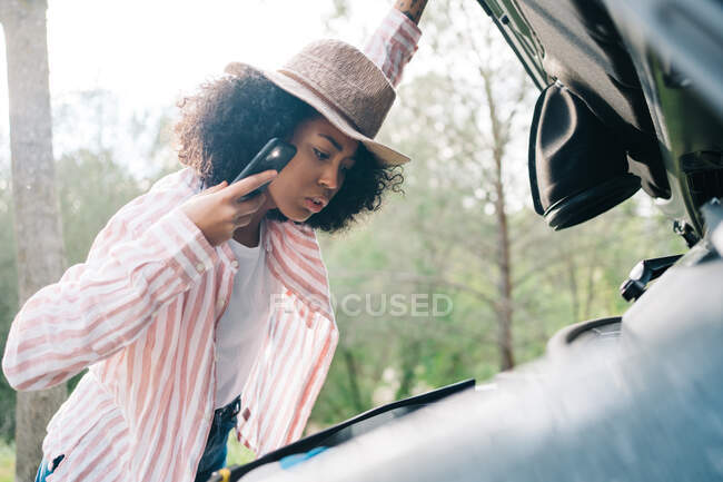 Side view of young African American female traveler holding smartphone with glowing flashlight while checking car engine as having problem during road trip through nature — Stock Photo