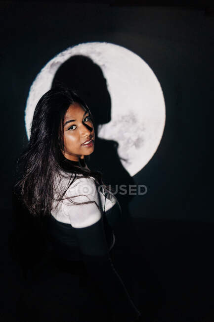 Vue latérale de la jeune femme hispanique regardant la caméra se reposer sous la projection de la lune la nuit — Photo de stock