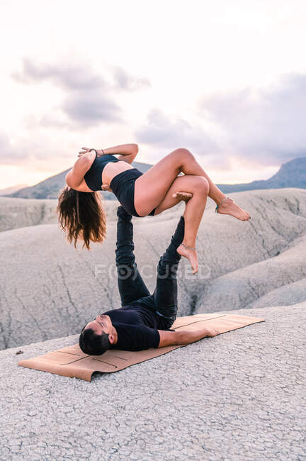 Mujer balanceándose con las manos de Namaste sobre las piernas del hombre mientras practican acroyoga juntos en las tierras altas - foto de stock