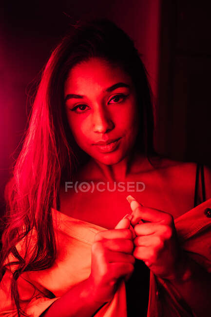 Sensual jovem hispânica fêmea étnica em lingerie olhando para a câmera enquanto descansa no terraço sob néon vermelho — Fotografia de Stock