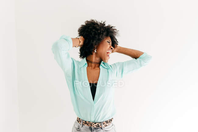 Playful young African American female with closed eyes in trendy outfit having fun touching afro hair on white background — Stock Photo