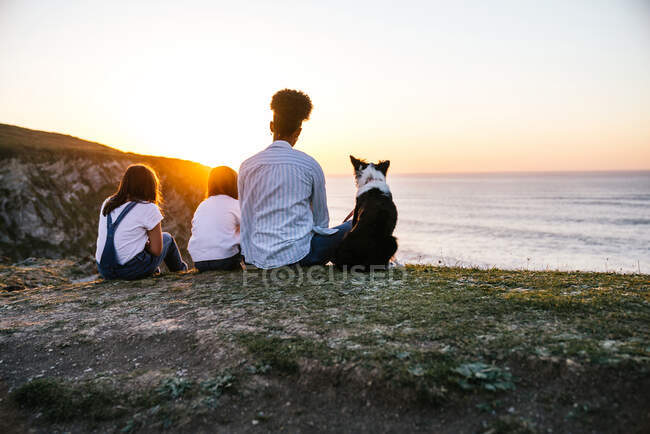 Back view of unrecognizable mother with children and Border Collie dog sitting on shore near sea and admiring sundown while spending evening together on beach — Stock Photo