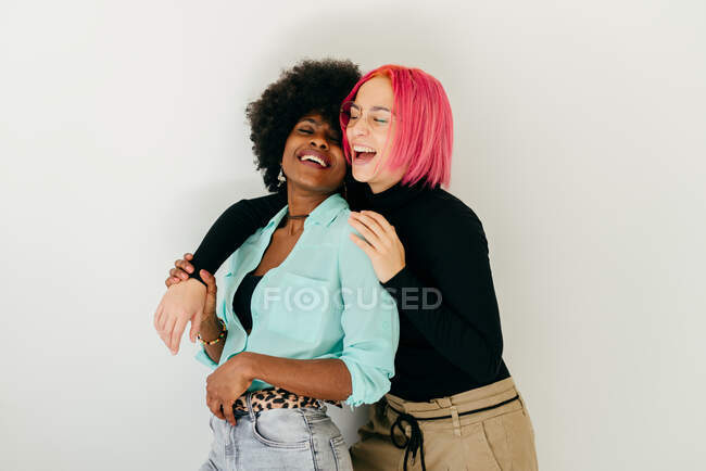 Веселая молодая розововолосая женщина, обнимающая позитивную афроамериканскую девушку в стильном наряде, веселясь вместе на белом фоне — стоковое фото