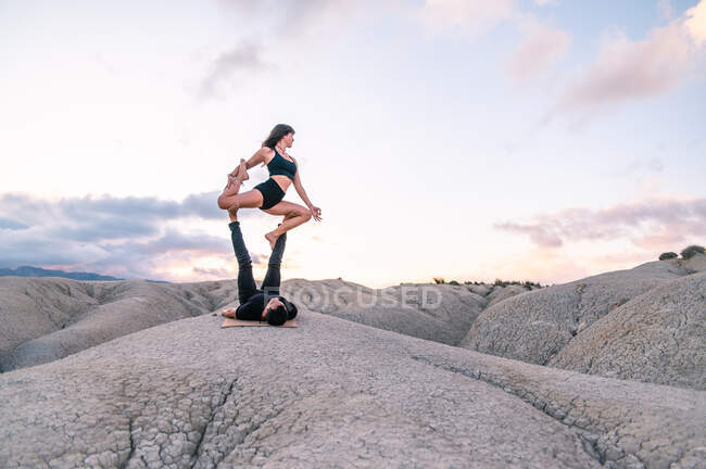 Serene woman balancing in King Pigeon pose on legs of man while practicing acroyoga in highlands — Stock Photo