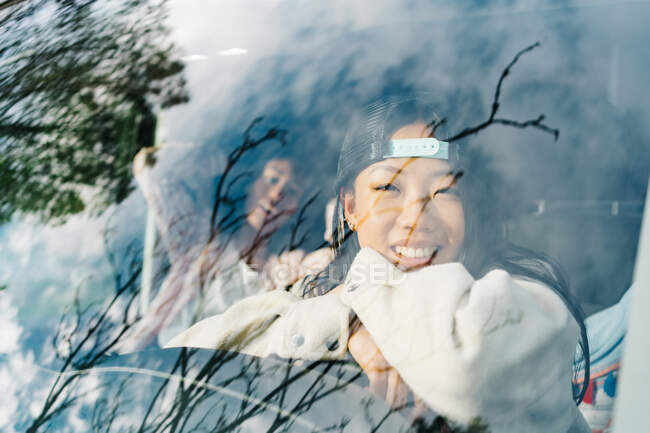 Through window of cheerful young Asian woman sitting on driver seat of camper vehicle and looking at camera while traveling with girlfriend through nature — Stock Photo