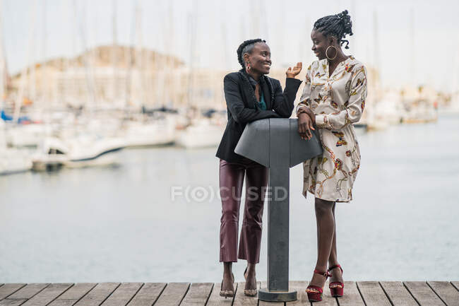 Модные хитроумные улыбчивые афроамериканские дамы проводят время вместе и выступают вместе в парке в яркий день — стоковое фото