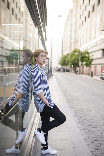 Vista lateral da moda modelo masculino asiático com cabelo loiro em pé perto de edifício de vidro na cidade — Fotografia de Stock