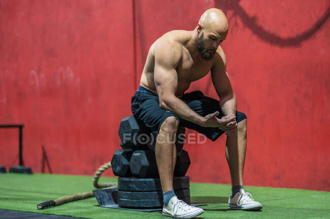 Вид сбоку измученного мускулистого мужчины, сидящего на гирях и отдыхающего во время функциональной тренировки в тренажерном зале — стоковое фото