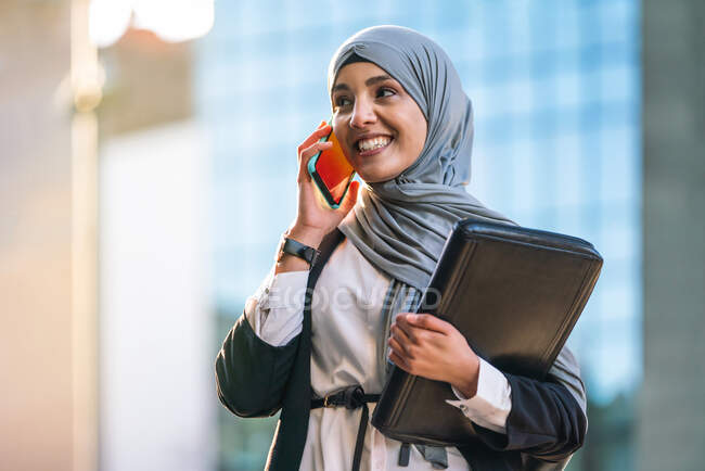 Contenuto Imprenditrice musulmana in hijab e con cartella in piedi in strada e parlando al telefono cellulare mentre discuteva del progetto aziendale — Foto stock