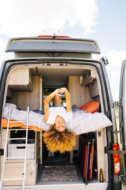 Positive schwarze Frau mit Afro-Frisur liegt kopfüber auf dem Bett im Wohnwagen und surft an sonnigen Sommertagen am Handy — Stockfoto