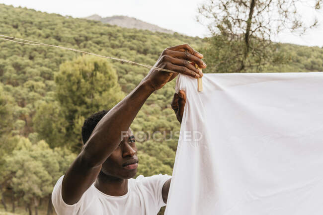 Hombre afroamericano enfocado colgando tela de algodón blanco en el tendedero en el campo en verano - foto de stock