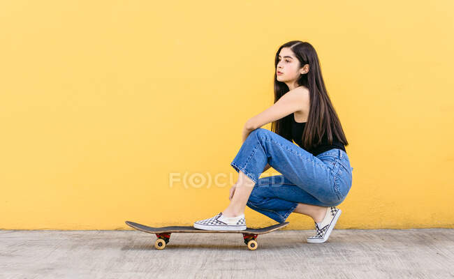 Vue latérale du jeune skateboarder féminin rêveur en tenue décontractée accroupi sur le trottoir tout en regardant loin — Photo de stock
