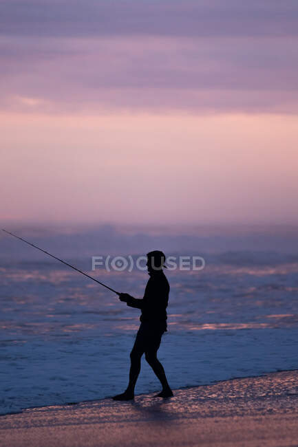 Silueta de un hombre pescando en la orilla del mar al atardecer - foto de stock