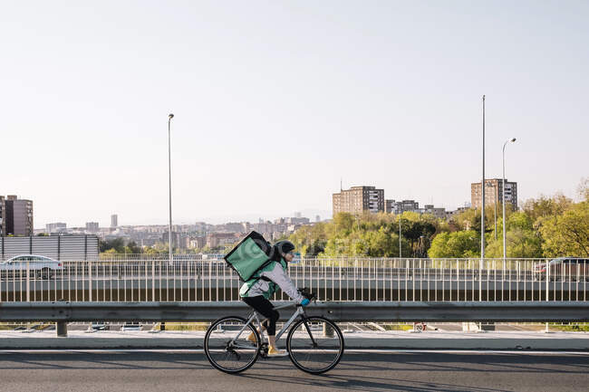 Side view of female courier with thermal bag riding bike on bridge while delivering food in city, copy space above — Stock Photo