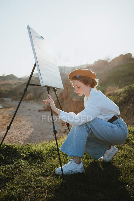 Вид збоку молодої стильної жінки-художниці в повсякденному одязі і береті, що встановлює мольберт на трав'янистій місцевості над піщаним пляжем хвилястого океану в сонячний день — стокове фото