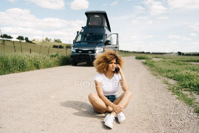 Worried young African American female sitting with curly hair talking on mobile phone while asking for help from repair service after accident with camper van in countryside — Stock Photo